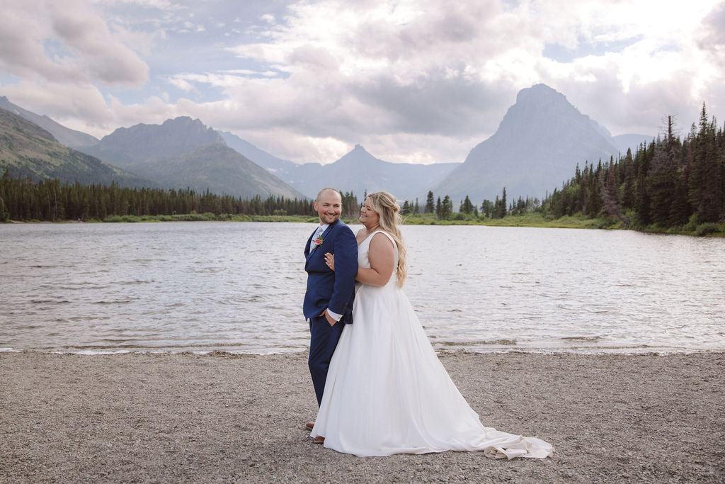 A bride and groom stand together by a lake, with mountains and trees in the background under a cloudy sky for a wedding at Glacier national park