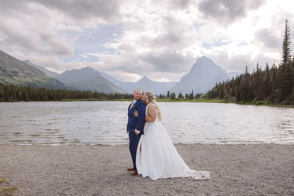 A bride and groom stand together by a lake, with mountains and trees in the background under a cloudy sky for a wedding at Glacier national park