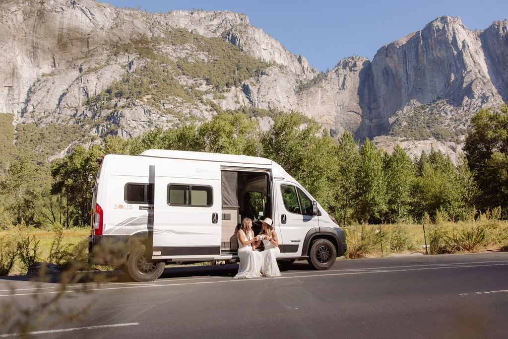Two women in white dresses sit on the ground next to a white camper van parked on a road, with large rocky mountains and trees in the background.