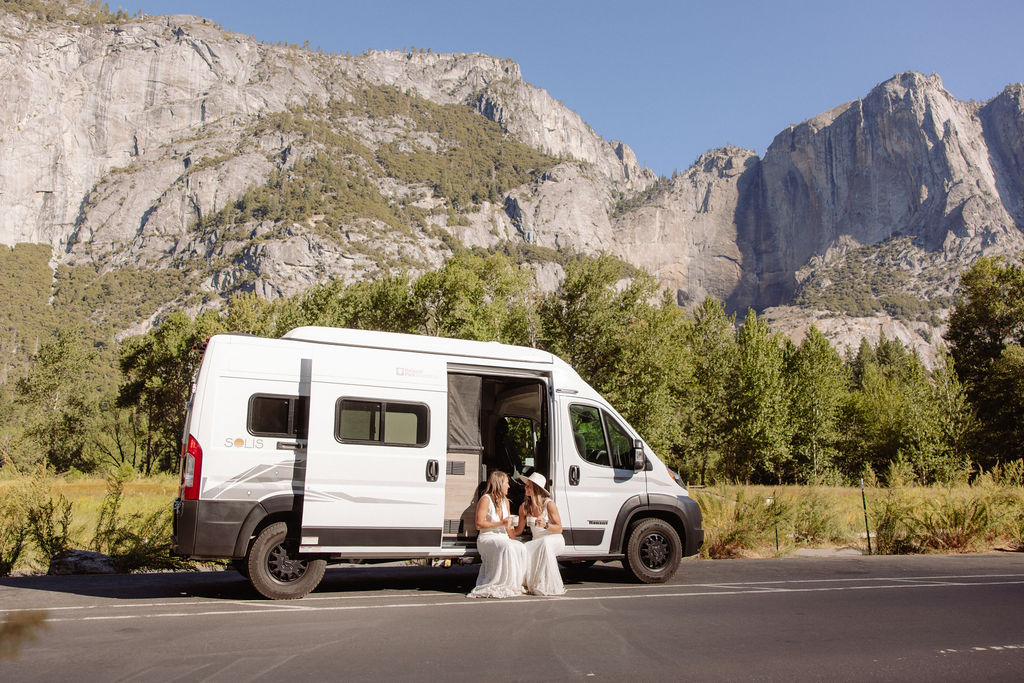 Two women in white dresses sit on the ground next to a white camper van parked on a road, with large rocky mountains and trees in the background.