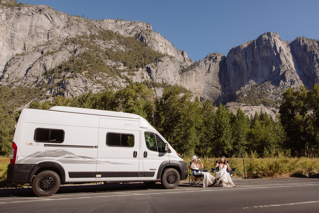 Two women in white dresses sit on the ground next to a white camper van parked on a road, with large rocky mountains and trees in the background.