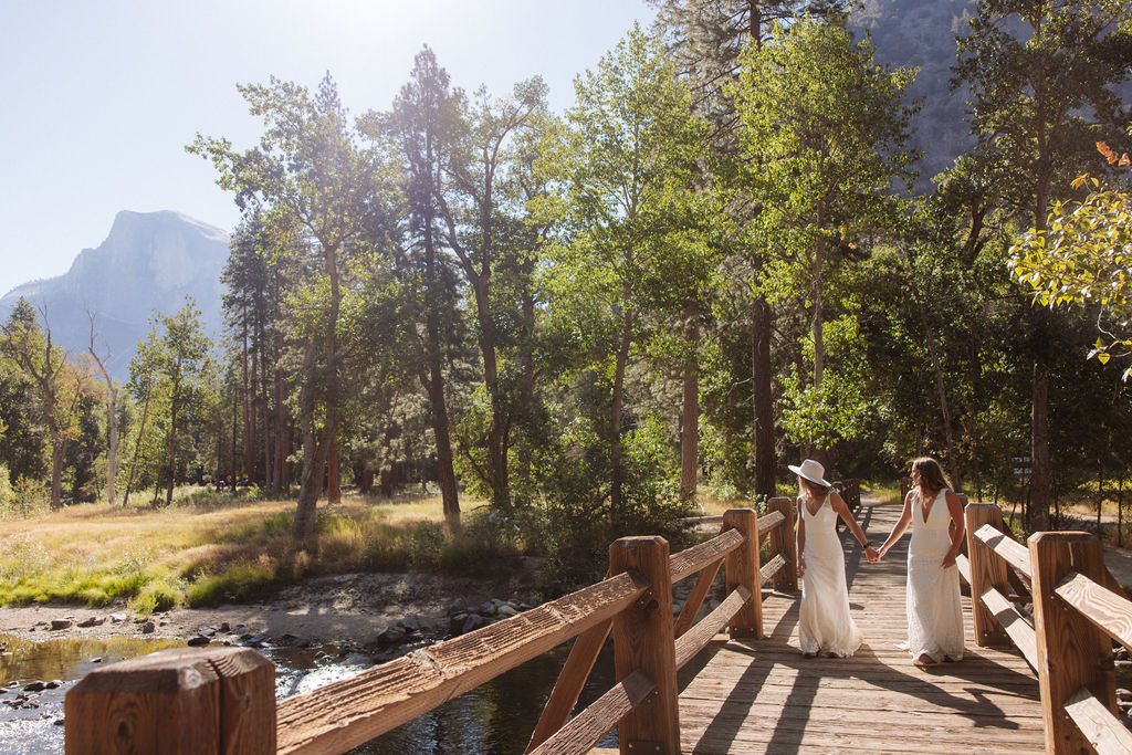 Two women in wedding dresses embrace and kiss on a wooden bridge in a forested area with mountains in the background.