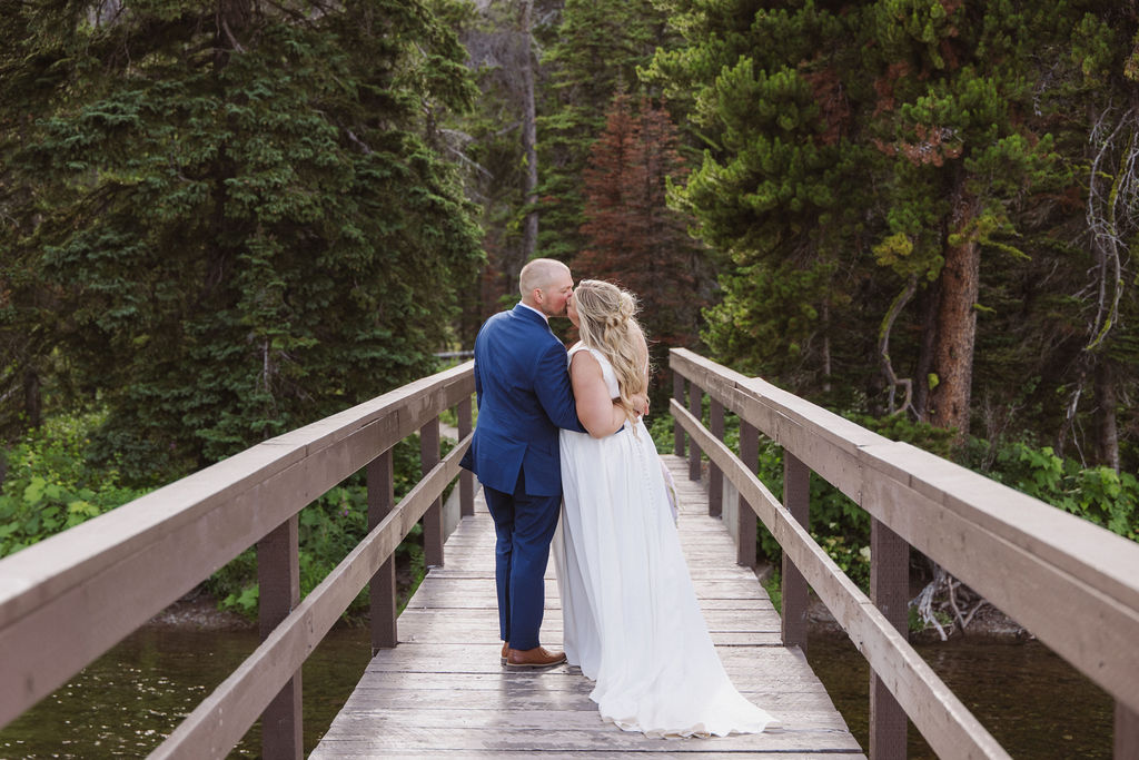 A couple in wedding attire stands on a wooden bridge surrounded by trees, facing away and embracing.