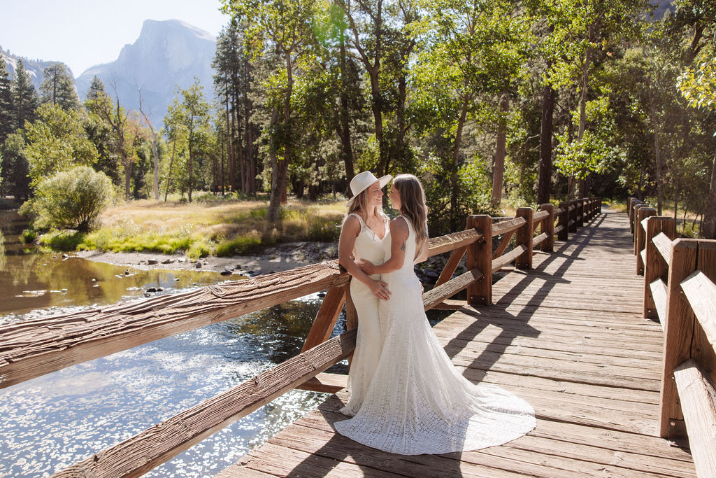 Two women in wedding dresses embrace and kiss on a wooden bridge in a forested area with mountains in the background.