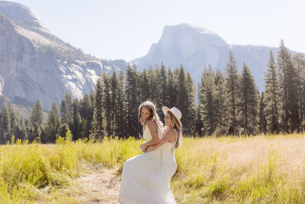 Two women in white dresses, one wearing a hat, stand together in a grassy field with mountains and pine trees in the background.
