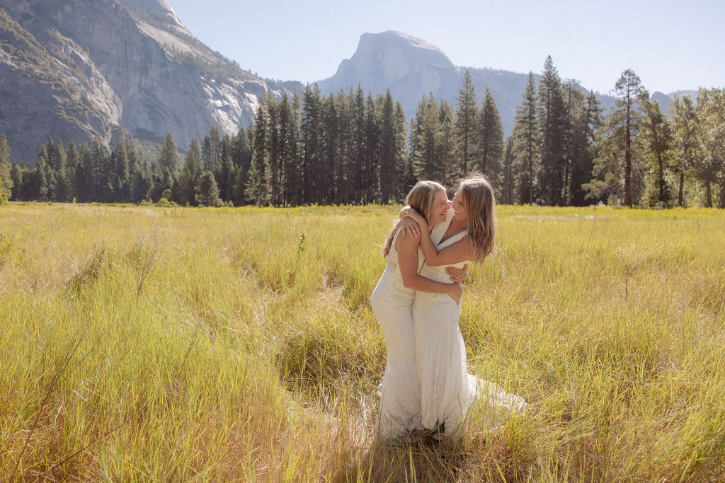 Two women in white dresses, one wearing a hat, stand together in a grassy field with mountains and pine trees in the background.