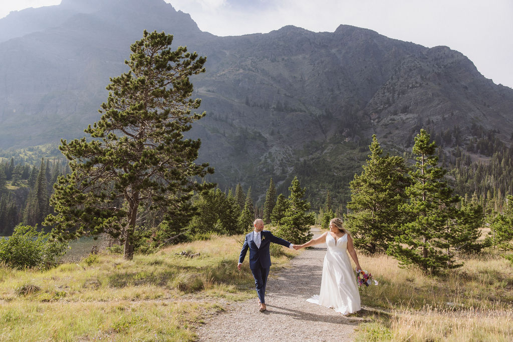 A couple in wedding attire stands on a wooden bridge surrounded by trees, facing away and embracing.