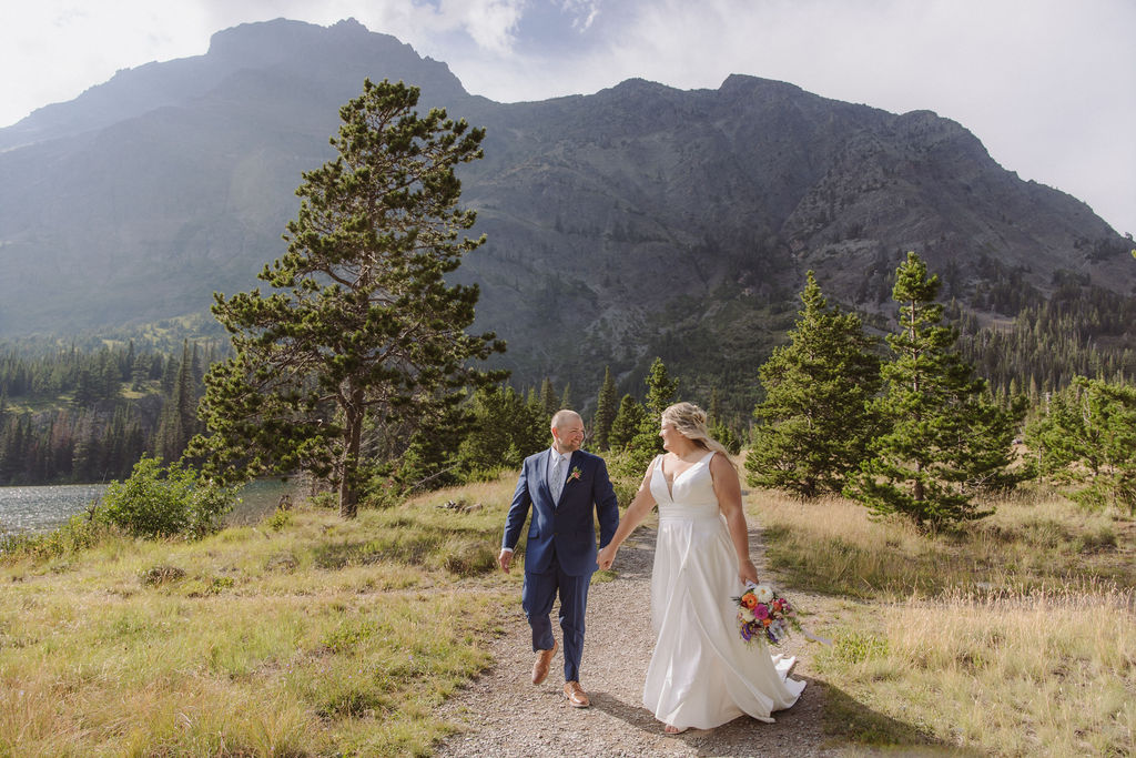 A couple in wedding attire stands on a wooden bridge surrounded by trees, facing away and embracing.
