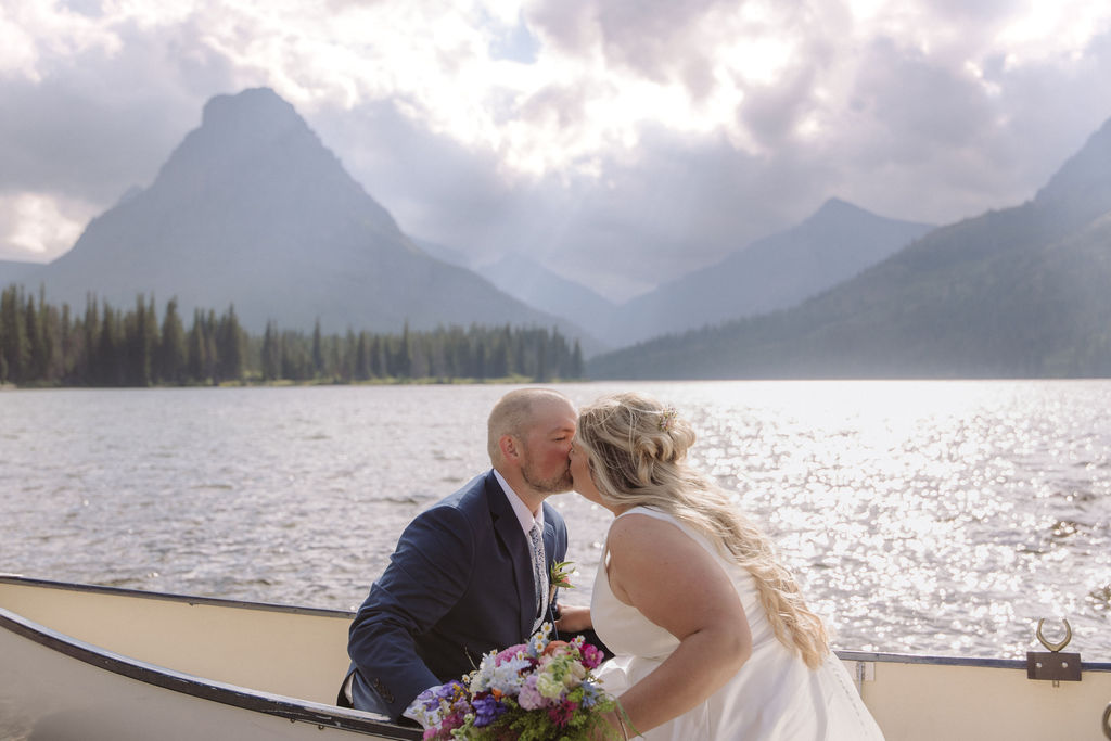A bride and groom kiss while sitting in a canoe on a lake, with mountains and pine trees in the background under a partly cloudy sky.