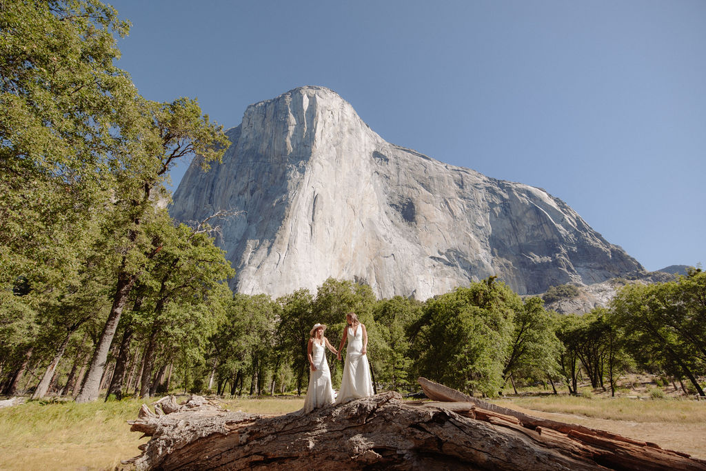 Two women in white dresses, one wearing a hat, stand together in a grassy field with mountains and pine trees in the background.