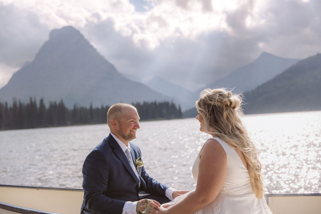 A bride and groom kiss while sitting in a canoe on a lake, with mountains and pine trees in the background under a partly cloudy sky.