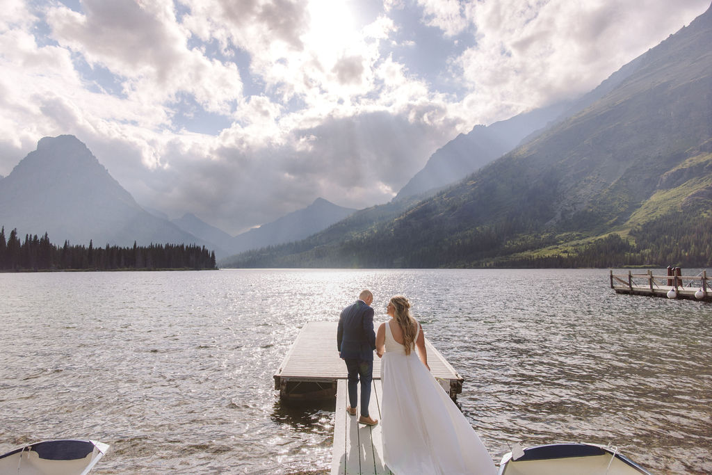 A couple in wedding attire walks on a dock extending into a lake, surrounded by mountains under a cloudy sky with sunlight streaming through.