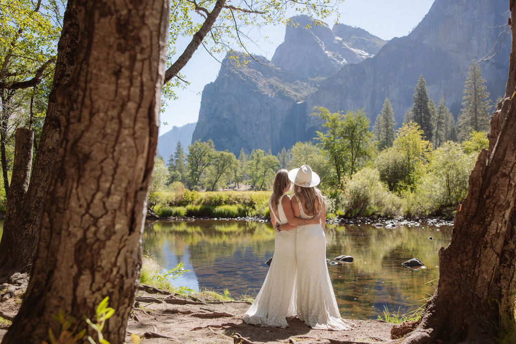 Two people in white dresses stand by a river, arm in arm, facing away, surrounded by trees and mountains on a sunny day.