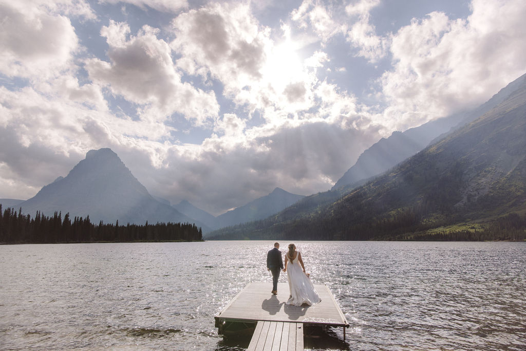 A couple in wedding attire walks on a dock extending into a lake, surrounded by mountains under a cloudy sky with sunlight streaming through.
