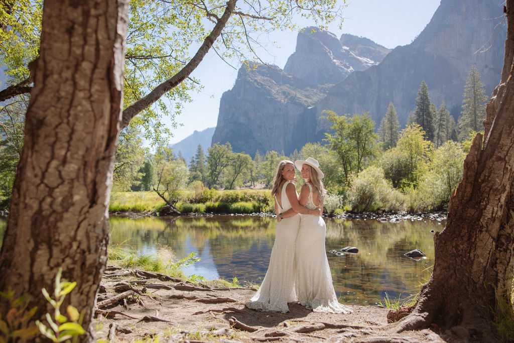 Two people in white dresses stand by a river, arm in arm, facing away, surrounded by trees and mountains on a sunny day.