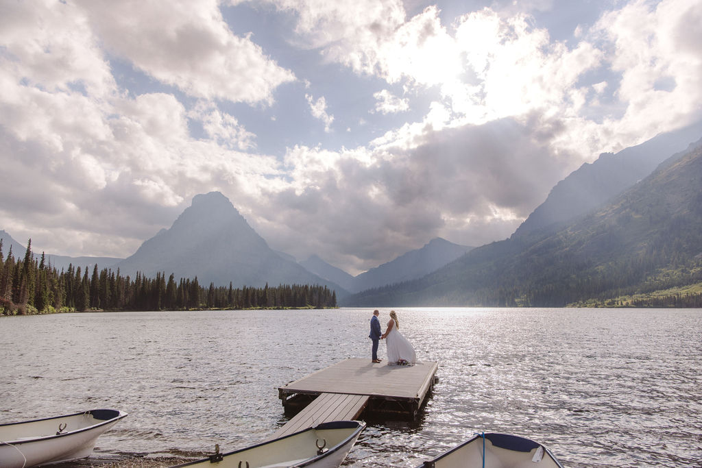 A couple in wedding attire walks on a dock extending into a lake, surrounded by mountains under a cloudy sky with sunlight streaming through.