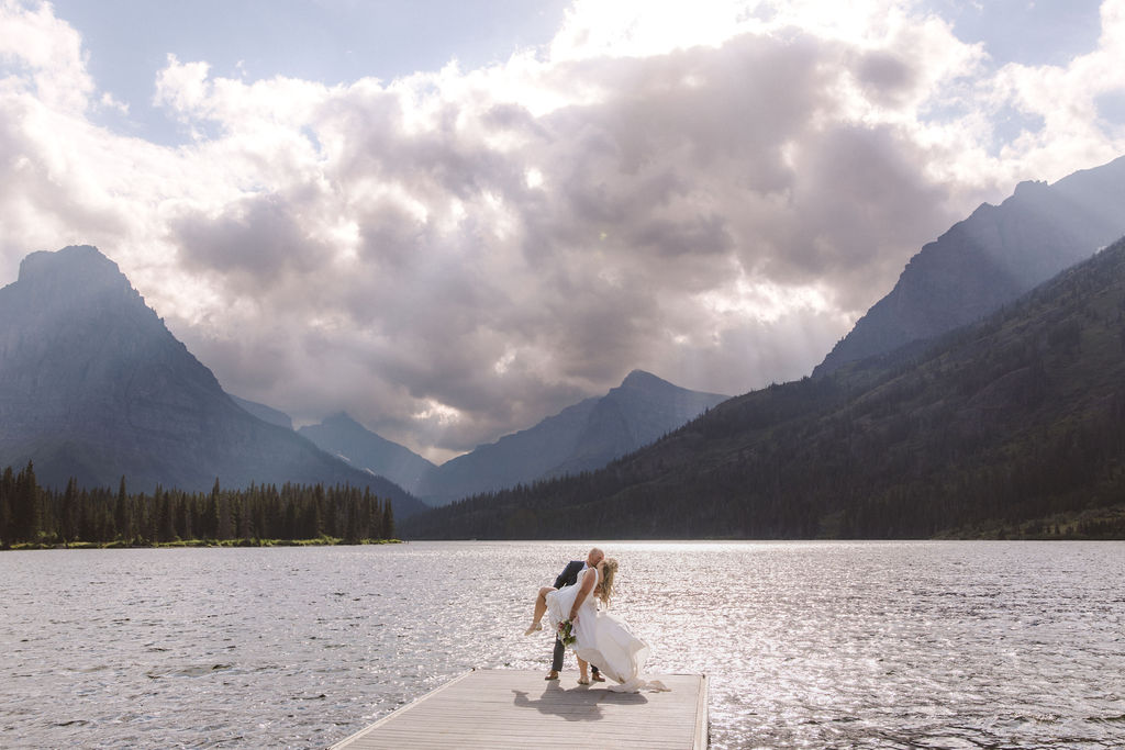 A couple in wedding attire walks on a dock extending into a lake, surrounded by mountains under a cloudy sky with sunlight streaming through.