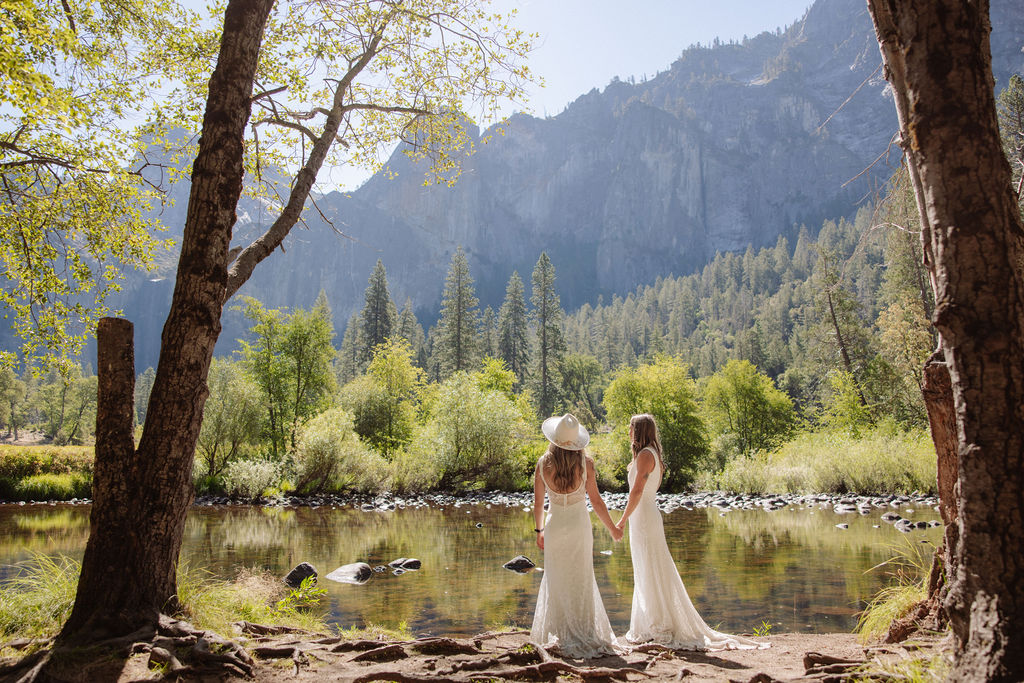 Two people in white dresses stand by a river, arm in arm, facing away, surrounded by trees and mountains on a sunny day.