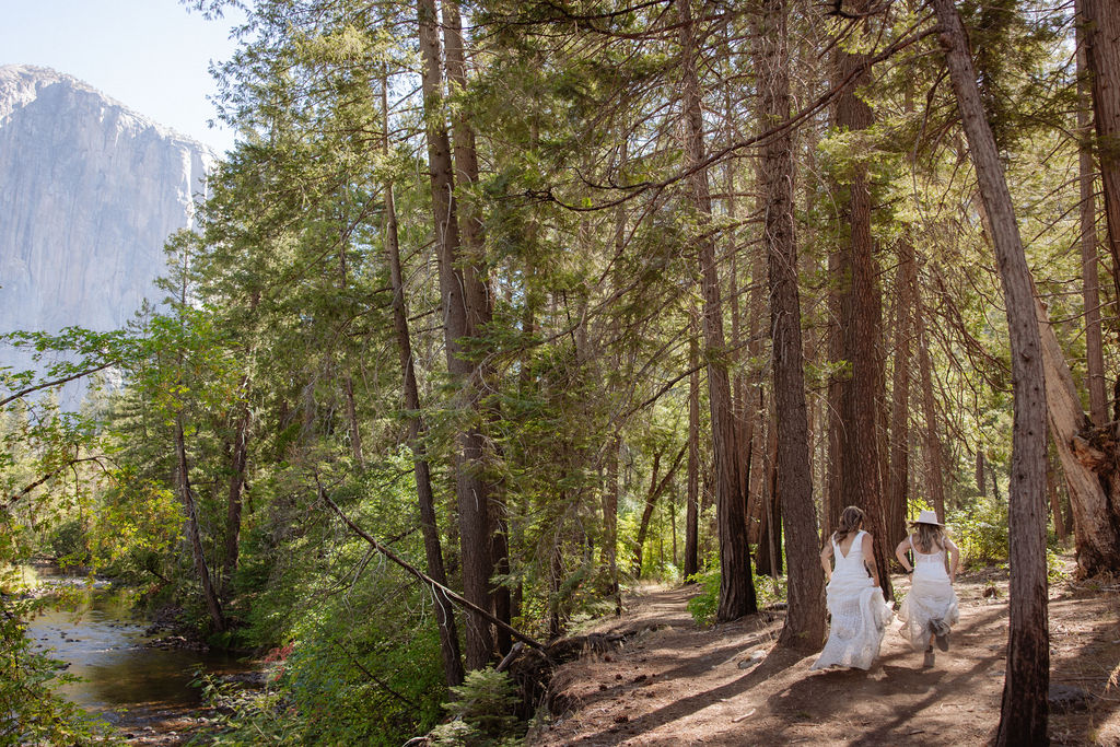 Two women in white dresses walk hand-in-hand along a dirt path in a forested area with a mountain and river in the background.