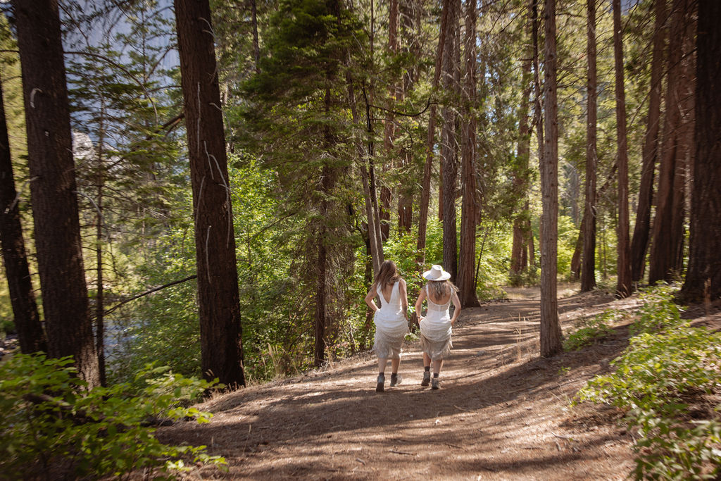 Two women in white dresses walk hand-in-hand along a dirt path in a forested area with a mountain and river in the background.
