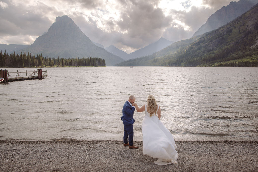 A bride and groom stand at the edge of a lake with mountains and cloudy sky in the background.