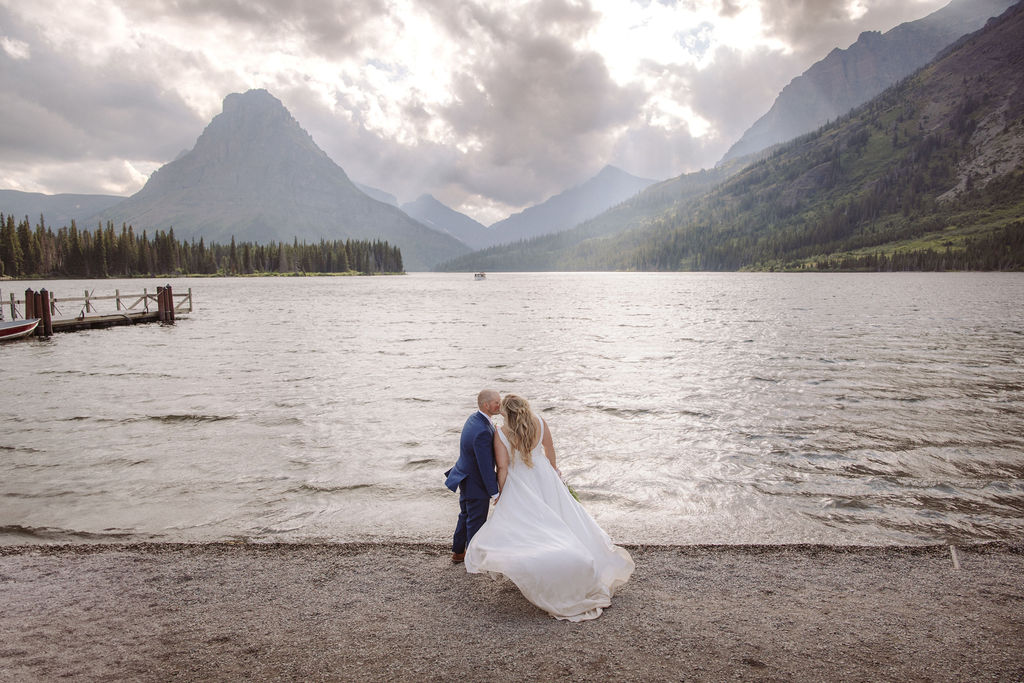 A bride and groom stand at the edge of a lake with mountains and cloudy sky in the background.