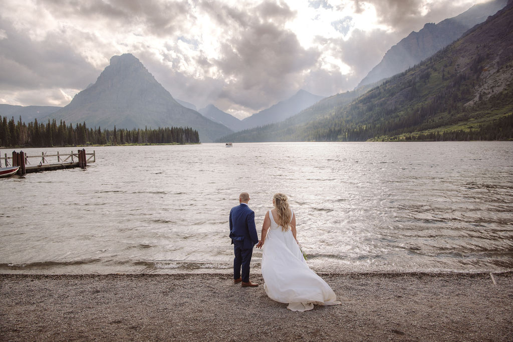 A bride and groom stand at the edge of a lake with mountains and cloudy sky in the background.
