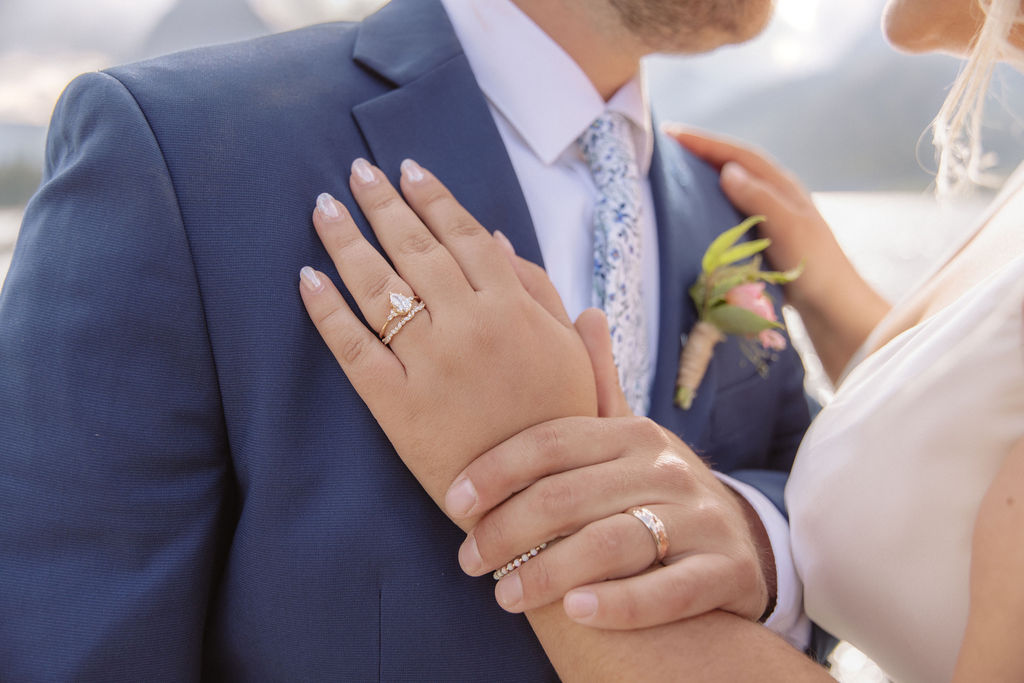 Close-up of a couple embracing outdoors, showing wedding rings on their hands; the groom wears a blue suit with a boutonniere, and the bride is in a white dress.