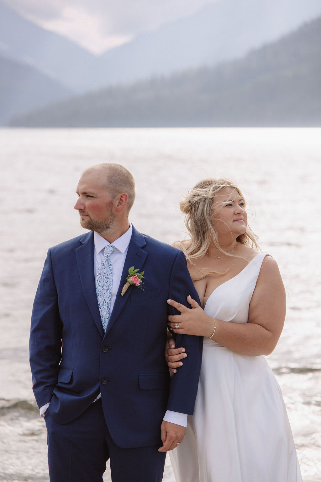 A bride in a white dress stands beside a groom in a blue suit by a lake, with mountains and water in the background. Both look in different directions.