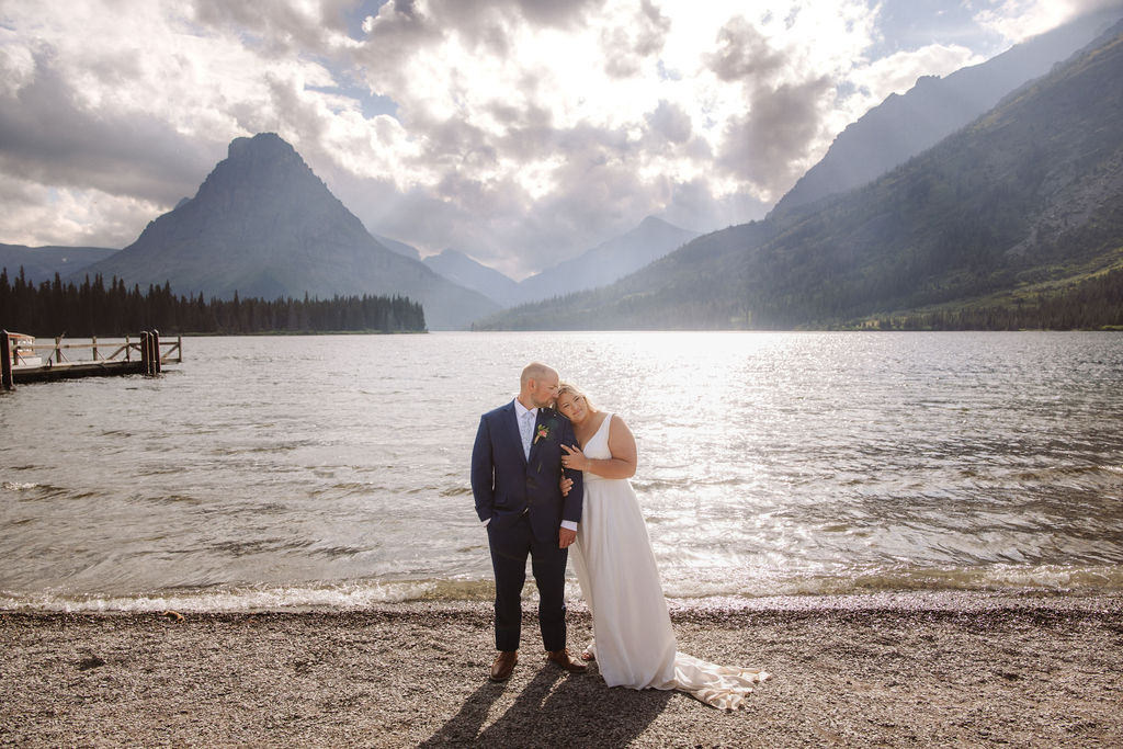 Close-up of a couple embracing outdoors, showing wedding rings on their hands; the groom wears a blue suit with a boutonniere, and the bride is in a white dress.