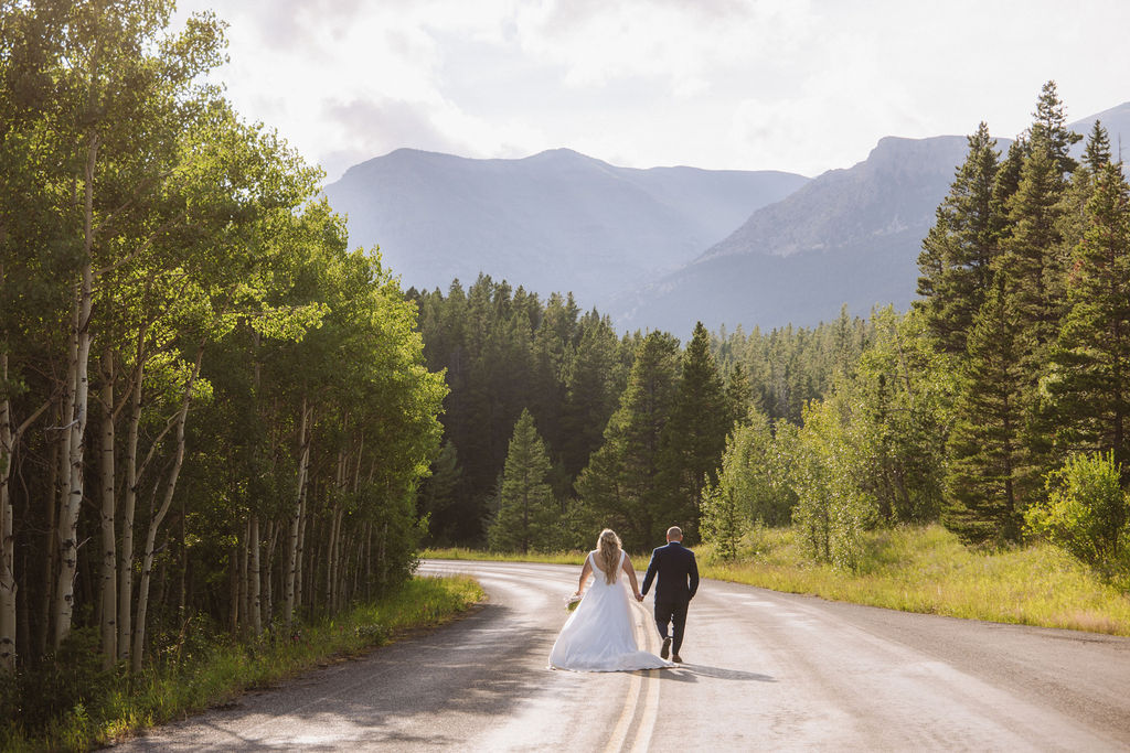 A couple in wedding attire holds hands while walking on a sunlit road surrounded by trees and mountains.