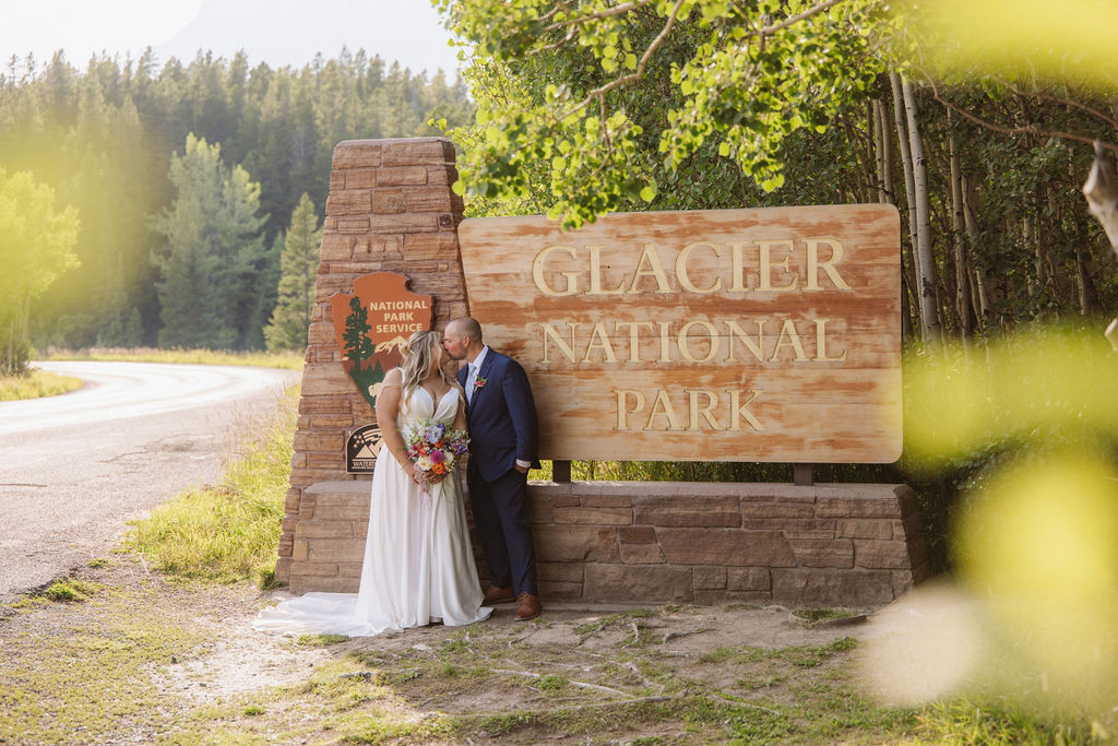 A bride and groom stand together in front of a large "Glacier National Park" sign surrounded by trees and greenery.