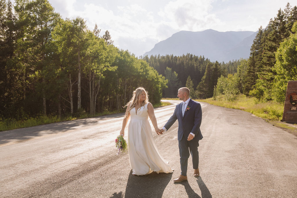 A couple in wedding attire holds hands while walking on a sunlit road surrounded by trees and mountains.