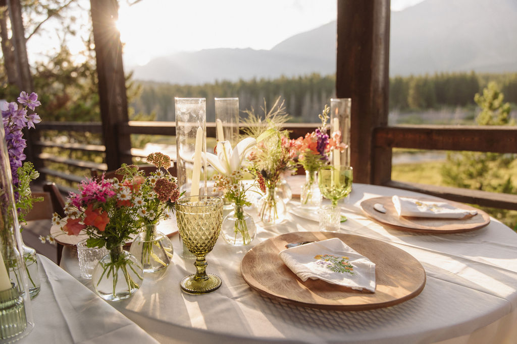 A round outdoor table is set with wooden plates, green glassware, napkins, and assorted small flower arrangements in glass vases, with a forest view in the background.