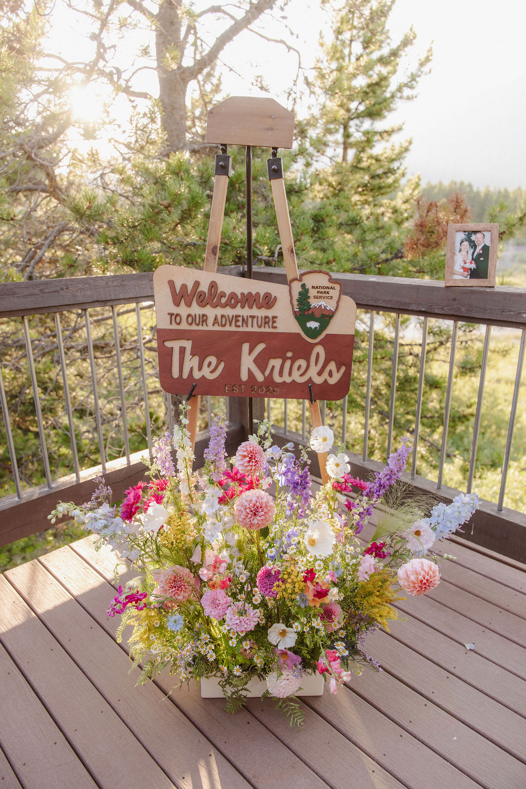 Wooden sign reads "Welcome to our adventure The Kriels," displayed above a basket of colorful flowers on a wooden deck, with trees and sunlight in the background.