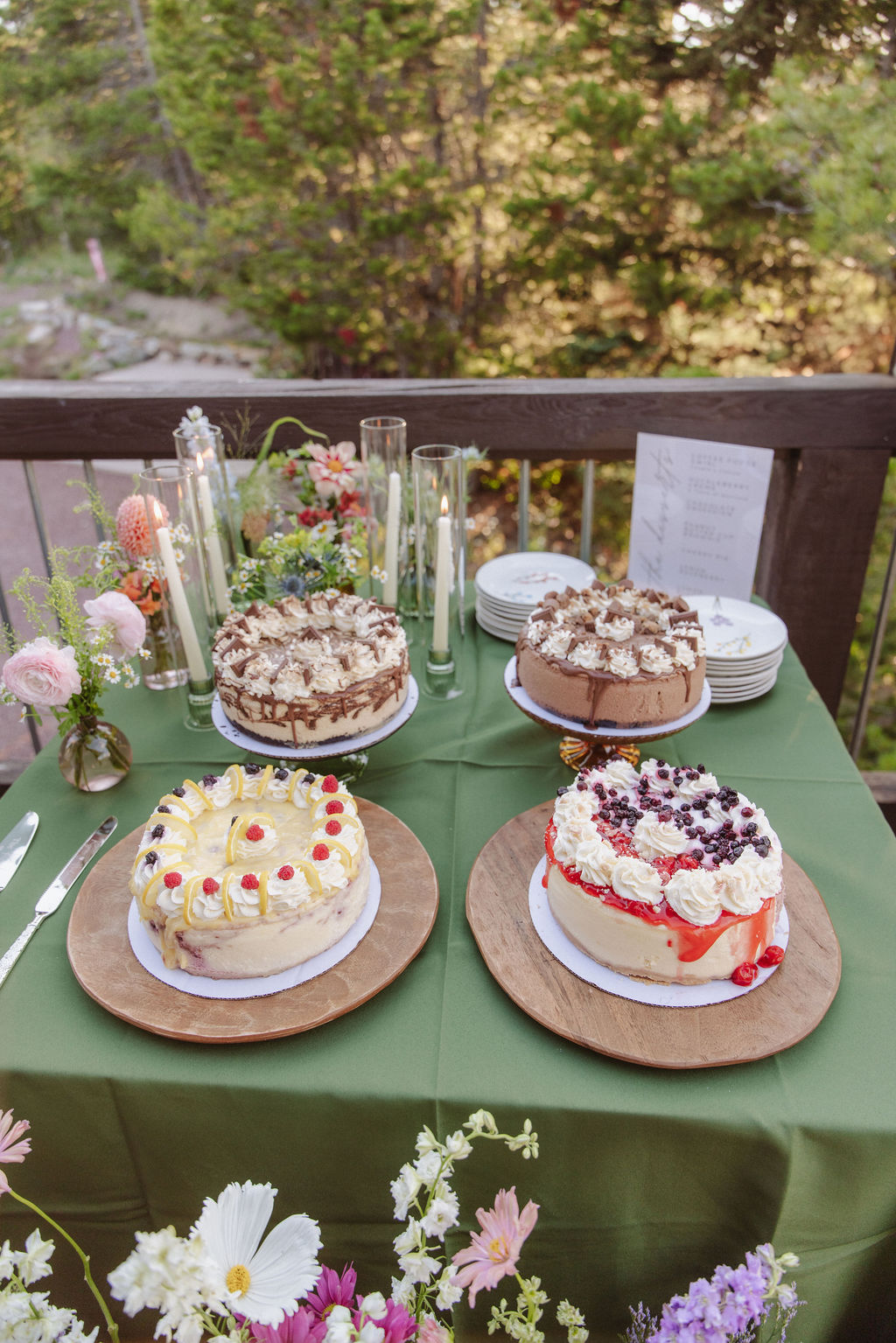 A green table with four decorated cakes on wooden trays, surrounded by plates, silverware, candles, and flower arrangements outdoors.