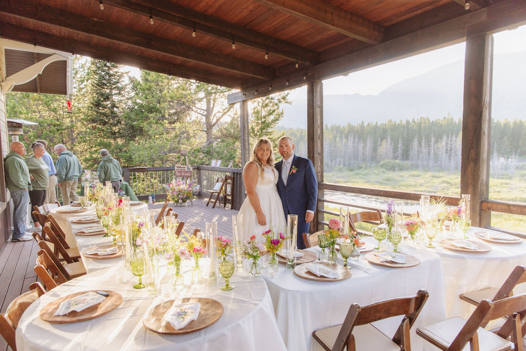 A bride and groom stand together on a decorated outdoor deck, surrounded by set dining tables with floral centerpieces and scenic mountain views in the background.