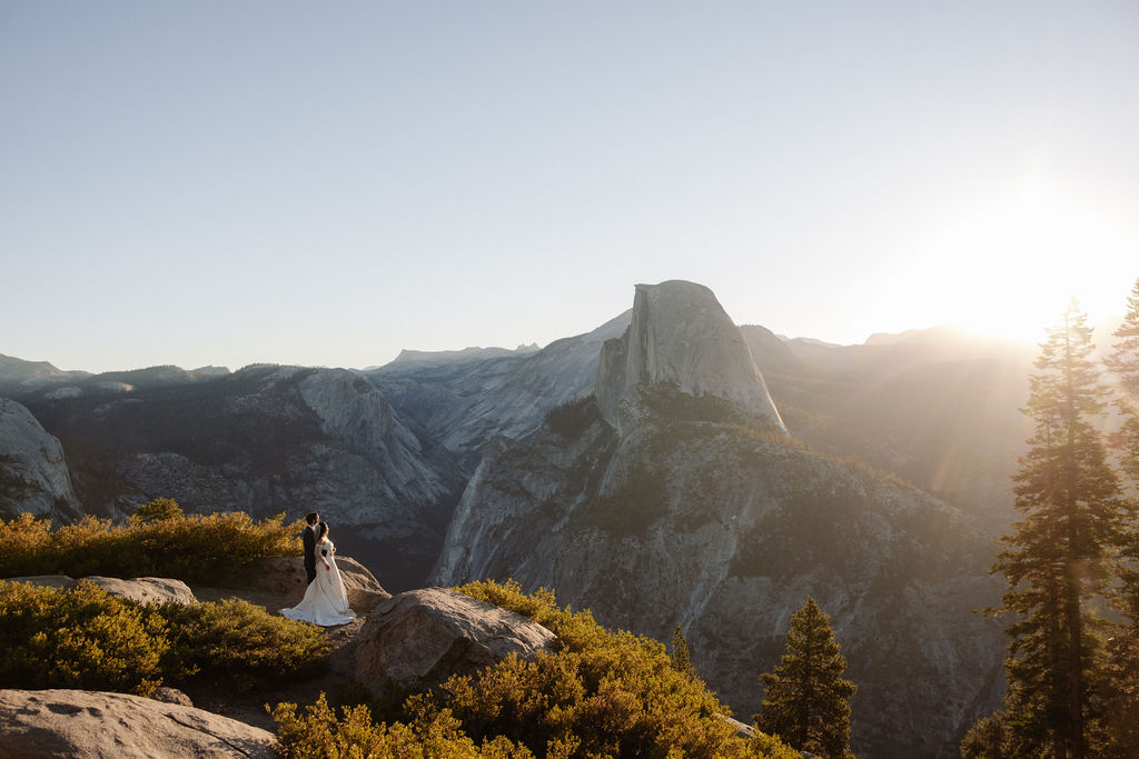 A couple in wedding attire stands on a rocky outcrop overlooking a mountainous landscape at sunset.