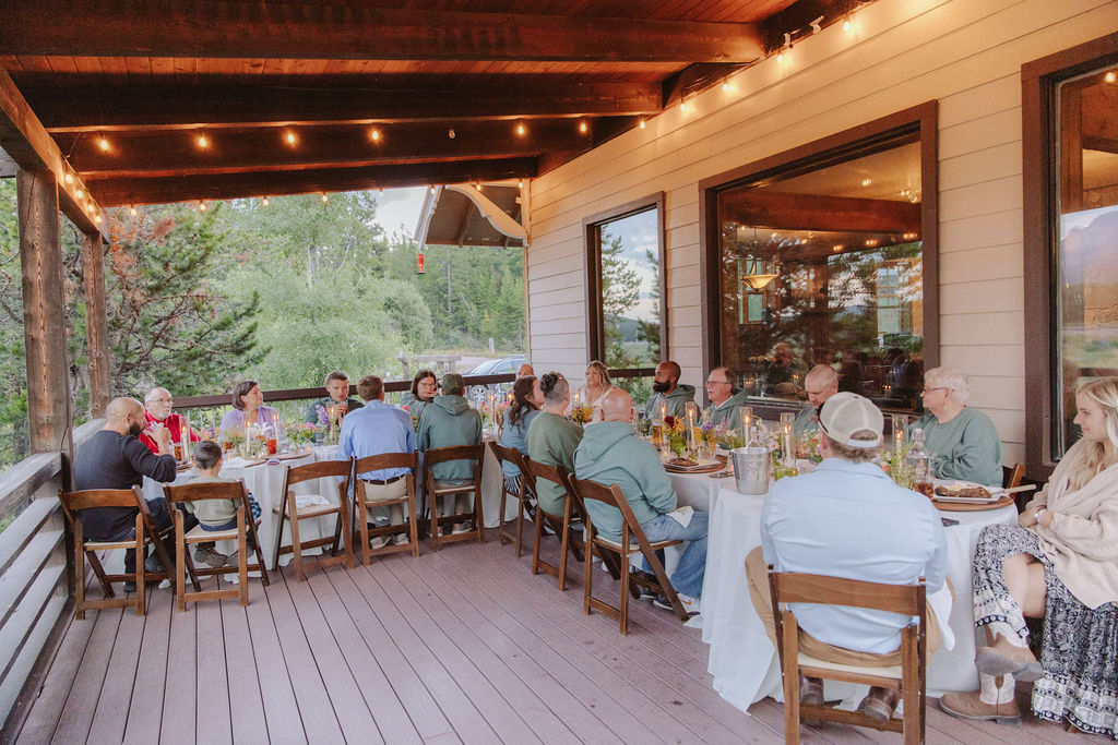 A bride and groom stand together on a decorated outdoor deck, surrounded by set dining tables with floral centerpieces and scenic mountain views in the background.