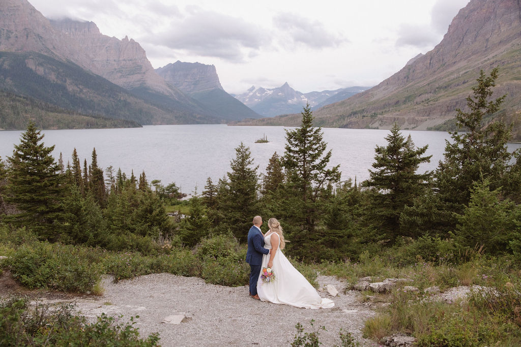A bride and groom stand together on a rocky path, surrounded by pine trees, overlooking a lake and mountains under a cloudy sky for a wedding at glacier national park