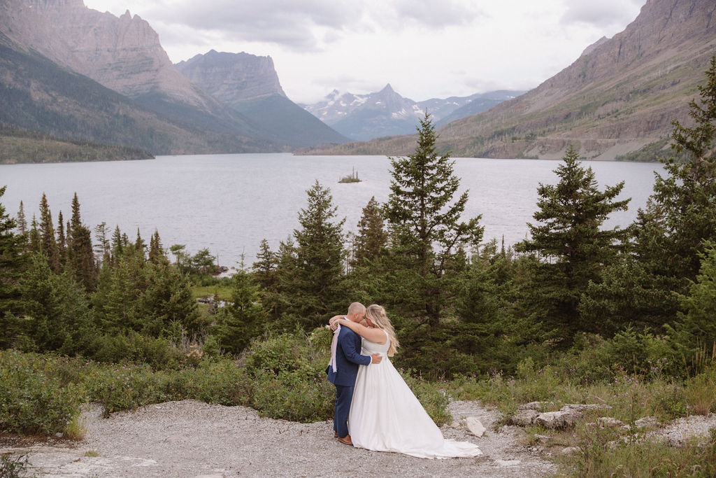 A bride and groom stand together on a rocky path, surrounded by pine trees, overlooking a lake and mountains under a cloudy sky for a wedding at glacier national park