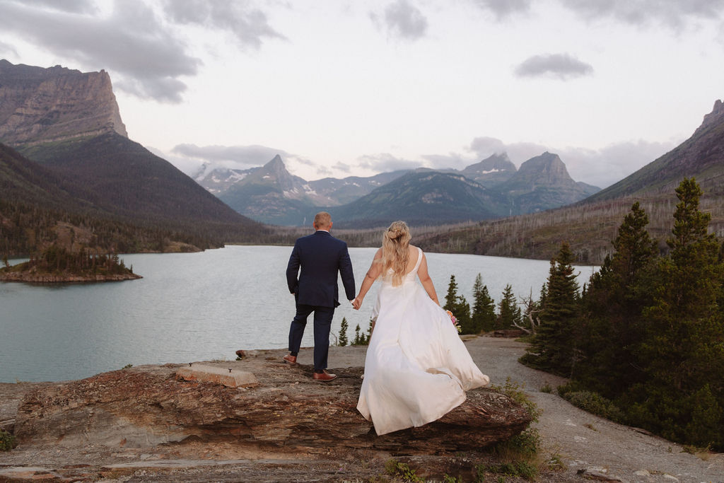A bride and groom stand together on a rocky path, surrounded by pine trees, overlooking a lake and mountains under a cloudy sky for a wedding at glacier national park  