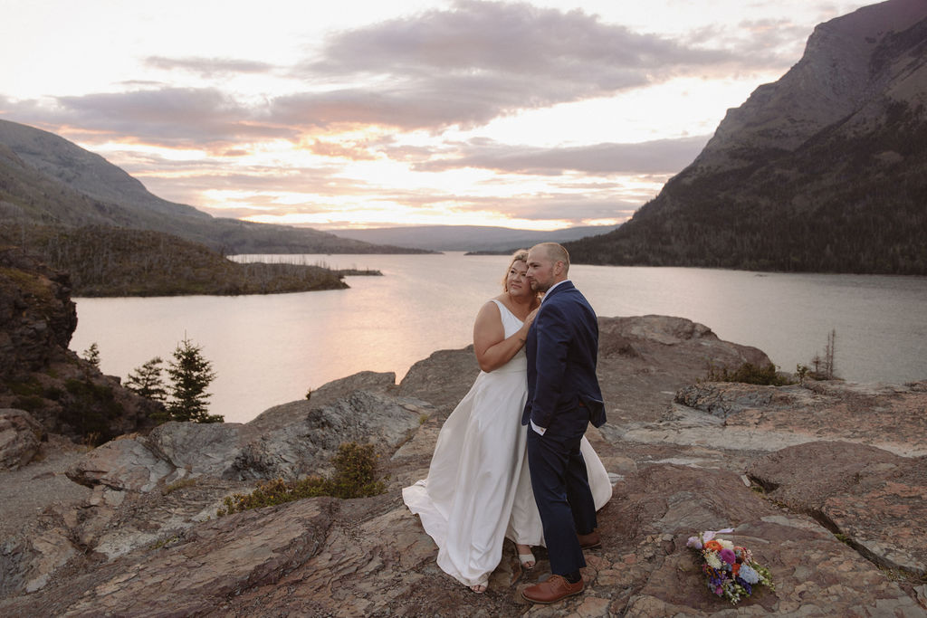 A bride and groom stand together on a rocky path, surrounded by pine trees, overlooking a lake and mountains under a cloudy sky for a wedding at glacier national park  