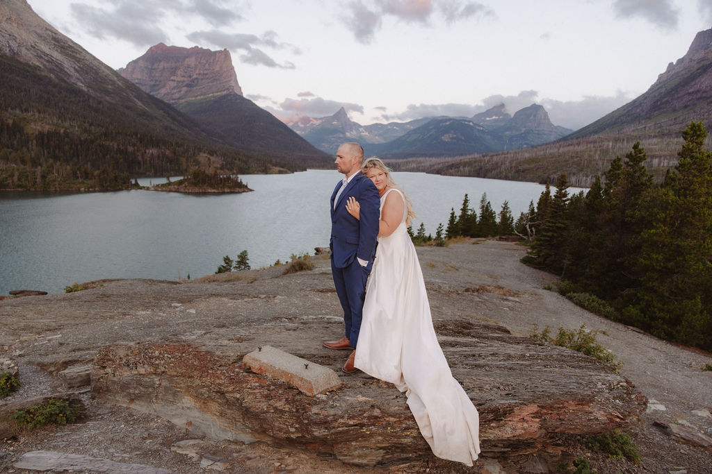 A bride and groom stand on rocky terrain overlooking a large lake and mountains, dressed in wedding attire, with trees and water in the background for a wedding at glacier national park