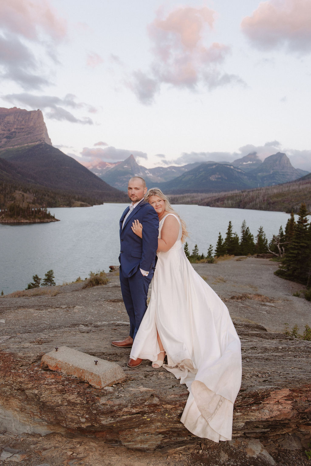 A bride and groom stand on a rocky overlook with a scenic mountain lake and cloudy sky in the background for a wedding at glacier national park