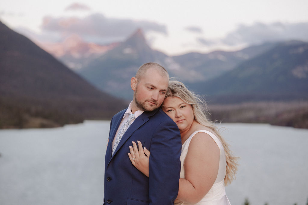 A couple in formal attire poses by a lake with mountains in the background during sunset for a wedding at glacier national park