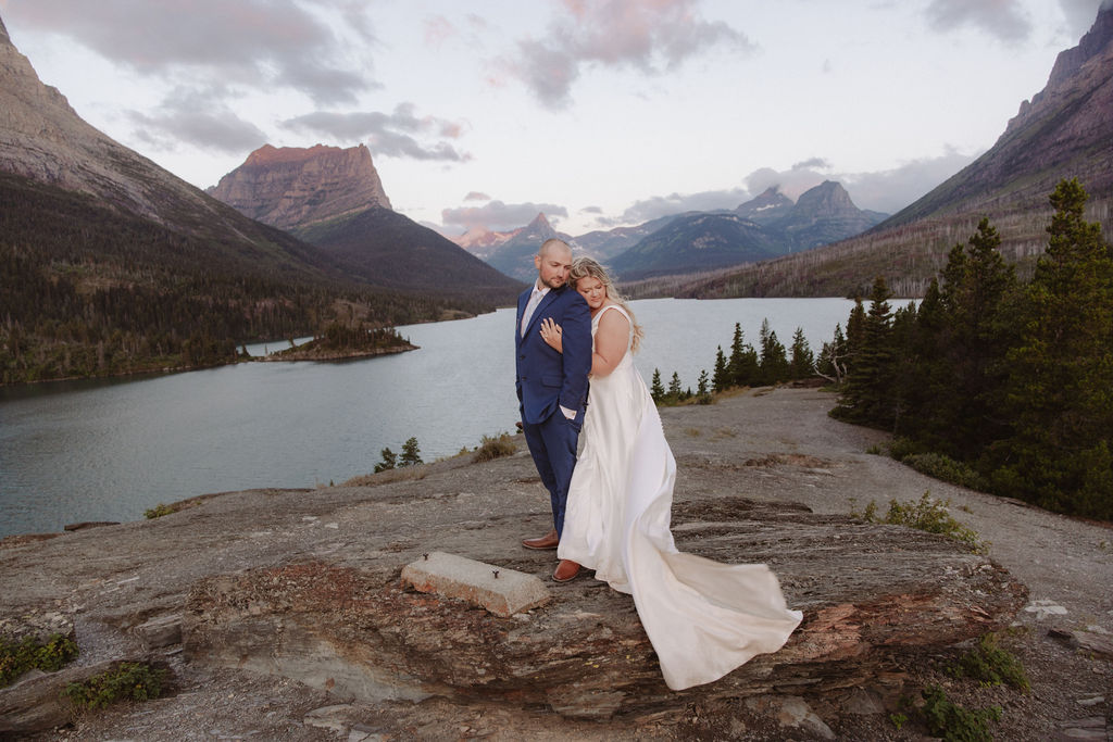 A couple in formal attire poses by a lake with mountains in the background during sunset or a wedding at glacier national park
