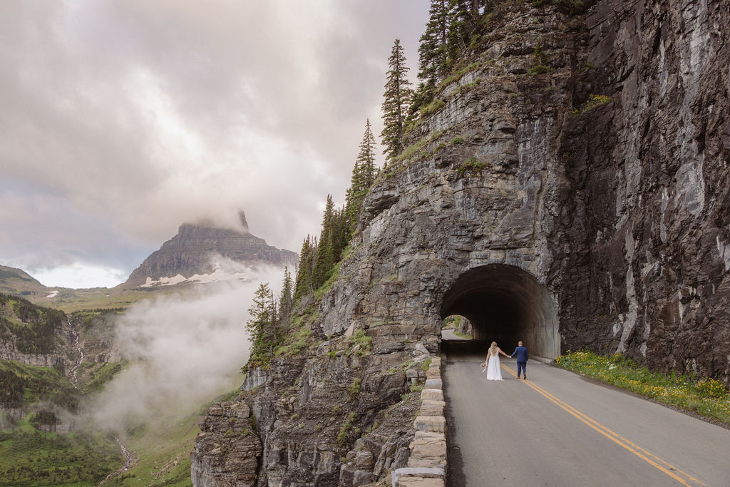 A bride and groom walk hand in hand on a mountain road beside a tunnel, with cliffs, mist, and a distant peak in the background or a wedding at glacier national park