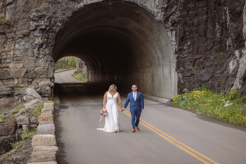 A bride and groom walk hand in hand on a mountain road beside a tunnel, with cliffs, mist, and a distant peak in the background or a wedding at glacier national park