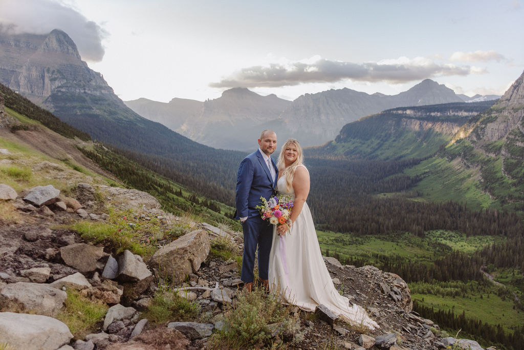 A bride and groom stand on rocky terrain overlooking a large lake and mountains, dressed in wedding attire, with trees and water in the background for a wedding at glacier national park